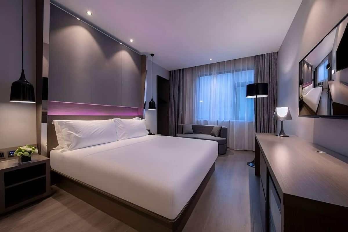 A modern hotel room with a large bed, a sofa, a desk, and a large window with curtains.