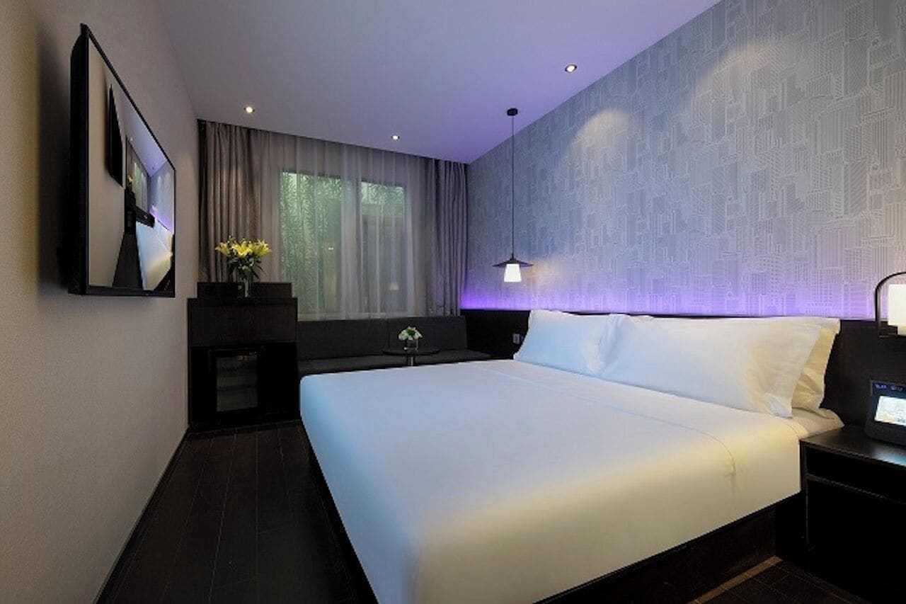 A modern hotel room with a large bed, a flat-screen TV mounted on the wall, a bedside table with a lamp, and a window with curtains.