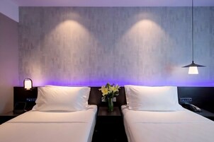 A hotel room with two beds, a nightstand with a lamp, a vase with flowers, and a wall with a geometric pattern.