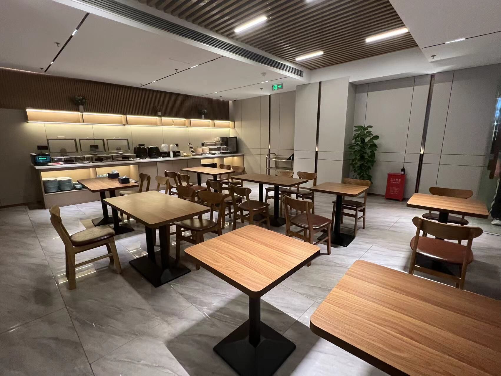 A modern restaurant interior with wooden tables and chairs, a kitchen area, and a potted plant.