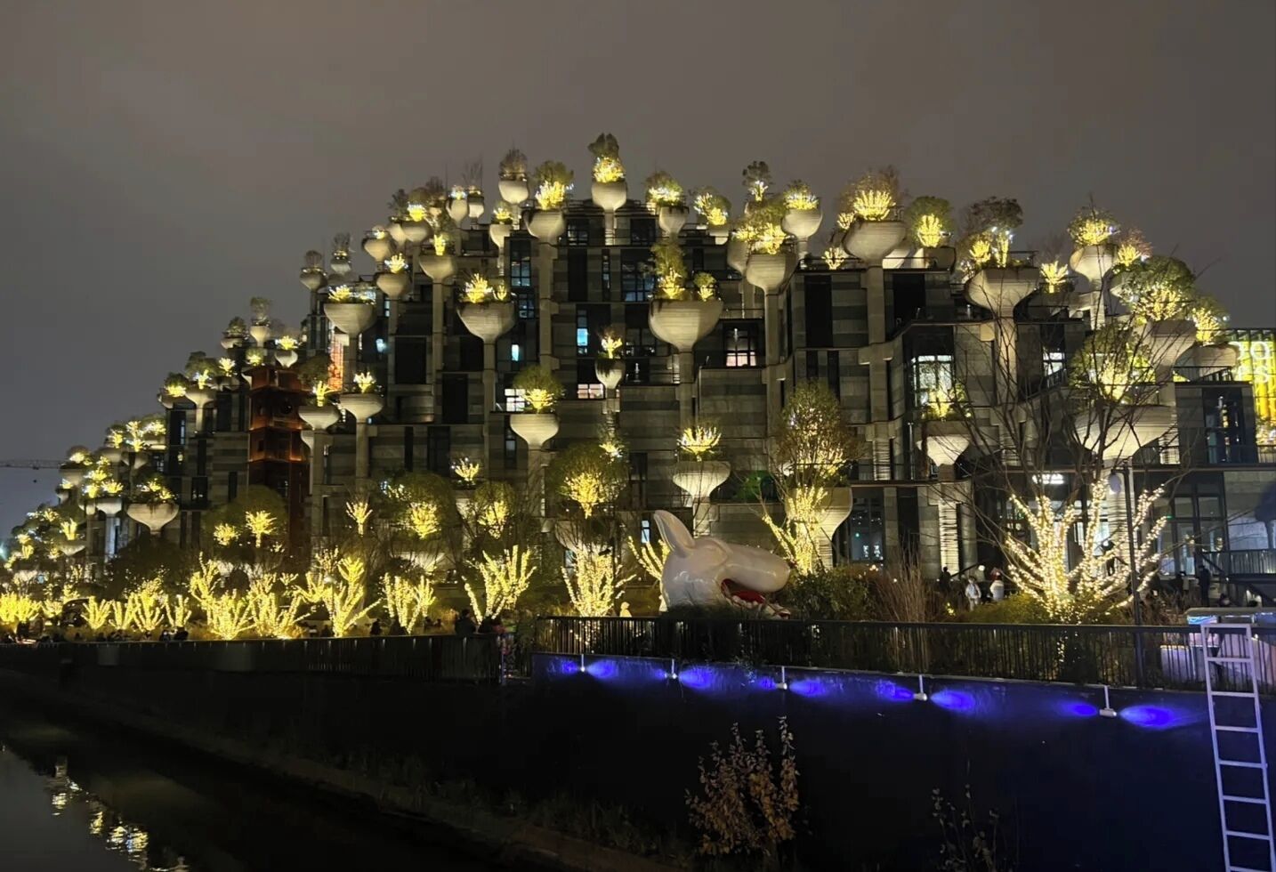 Un bâtiment à plusieurs étages, chaque étage étant orné de structures sphériques qui ressemblent à des arbres, et un plan d'eau au premier plan.
