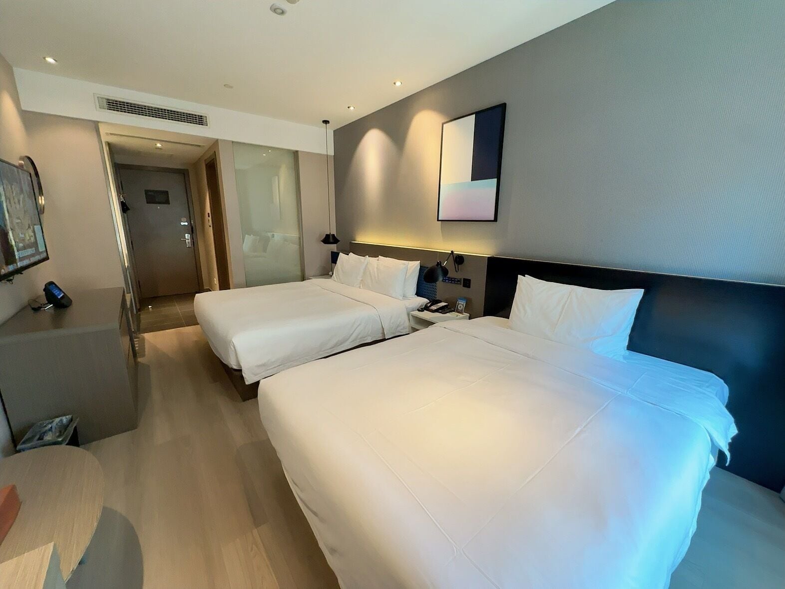 A hotel room with two beds, a desk, a chair, a TV, and a door leading to another room.