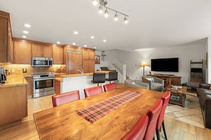 A modern kitchen with built-in appliances, a dining area with a wooden table and red chairs, a living room with a TV and sofa, and a staircase leading to an upper level.