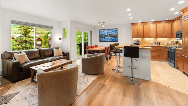 Een ruime woonkamer met een bank, salontafel en stoelen, een eethoek met tafel en stoelen, een keuken met houten kastjes en apparatuur, en grote ramen met uitzicht op groen.