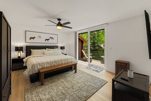 A bedroom with a bed, bedside lamps, a ceiling fan, a bench, a TV stand, and a sliding glass door leading to an outdoor deck.