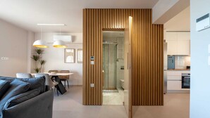 Een moderne woonkamer met een bank, een eettafel en hanglampen. Een houten scheidingswand leidt naar een badkamer met douche en toilet.