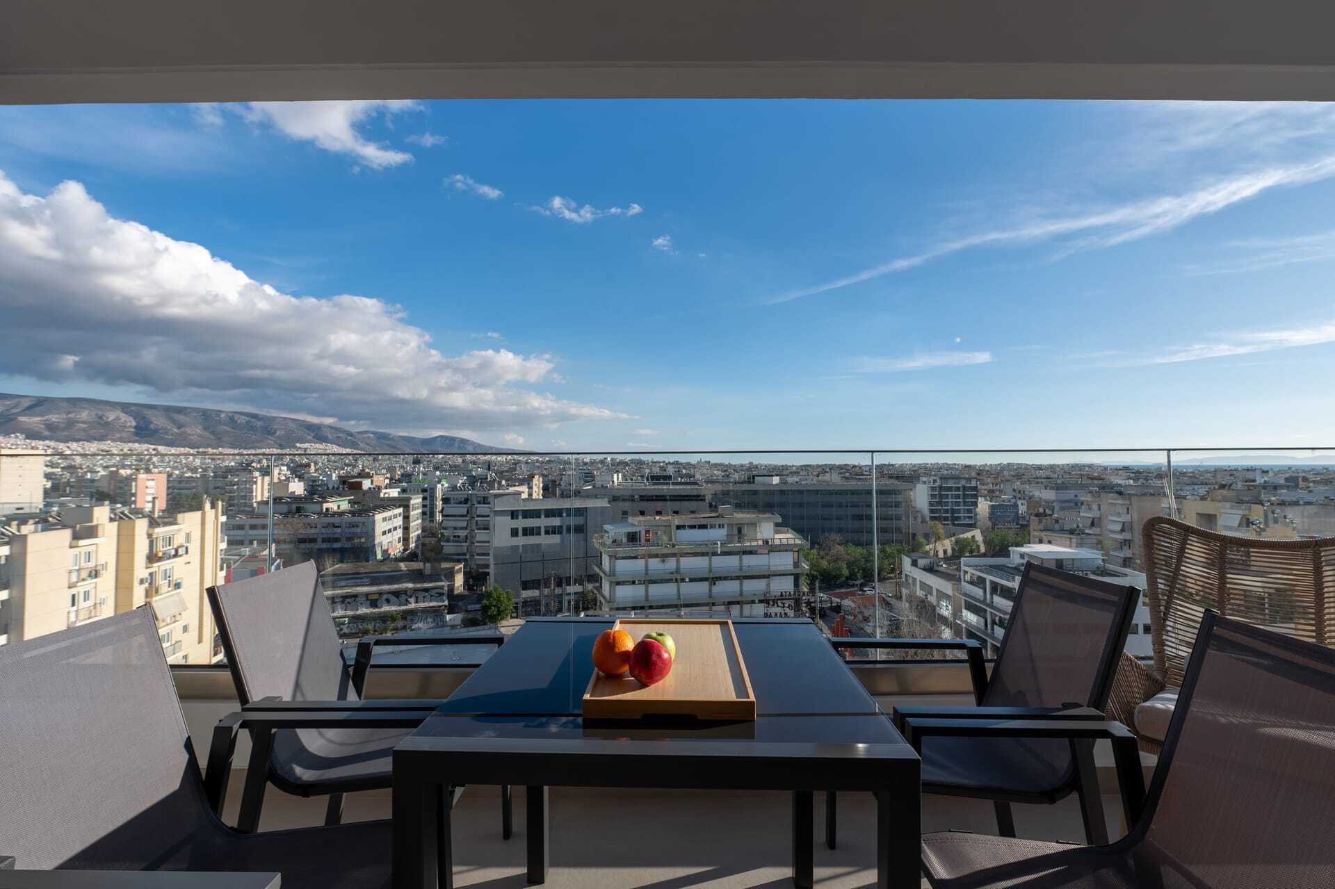 A balcony with a table set for two, overlooking a cityscape with mountains in the distance.