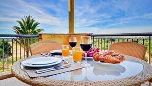 A round table with plates, cutlery, glasses of orange juice and red wine, and croissants on a balcony with a view of palm trees and the sky. - The Prime at The Residences Vilamoura (Loulé)
