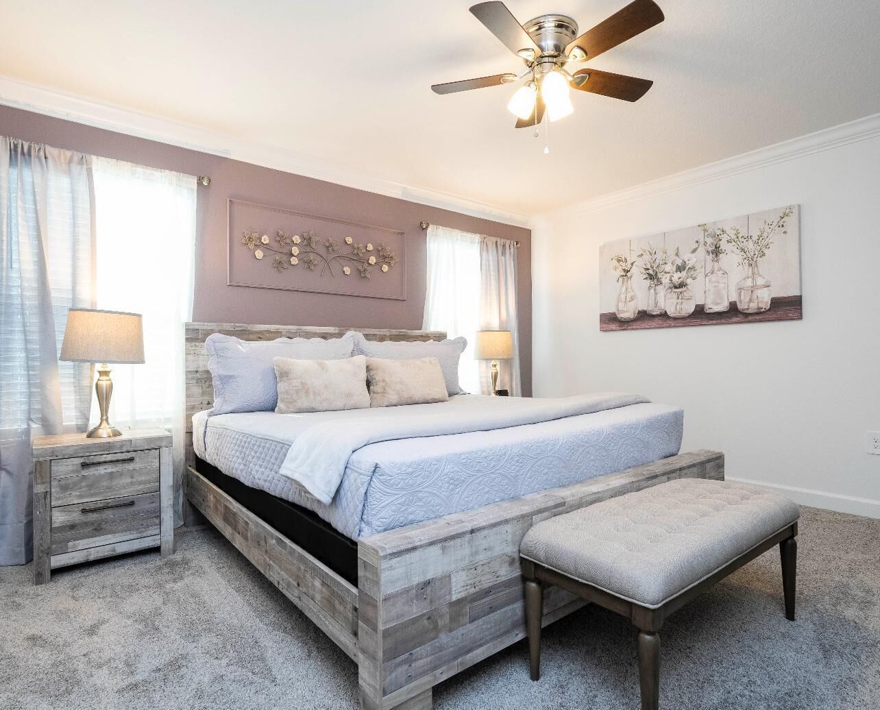 A bedroom with a wooden bed frame, a bench, a ceiling fan, and a painting on the wall.