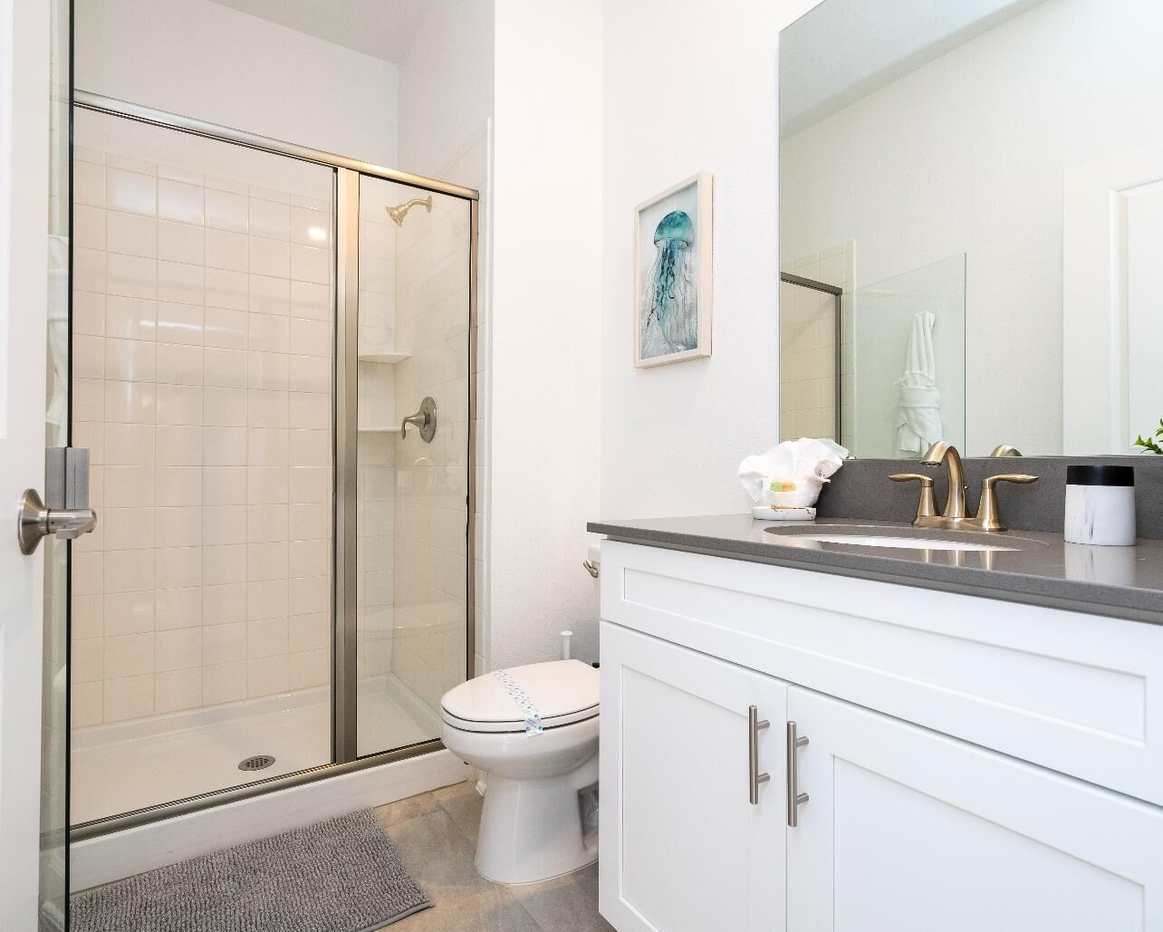 A bathroom with a glass-enclosed shower, a toilet, a sink with a mirror, and a framed artwork on the wall.