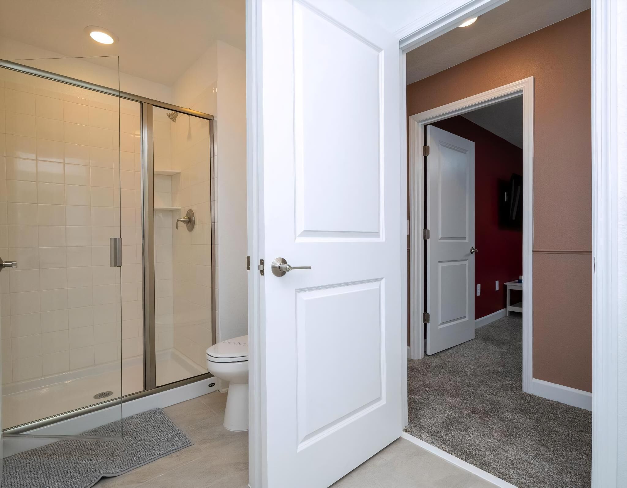 A bathroom with a glass shower enclosure, a toilet, and a white door leading to another room.