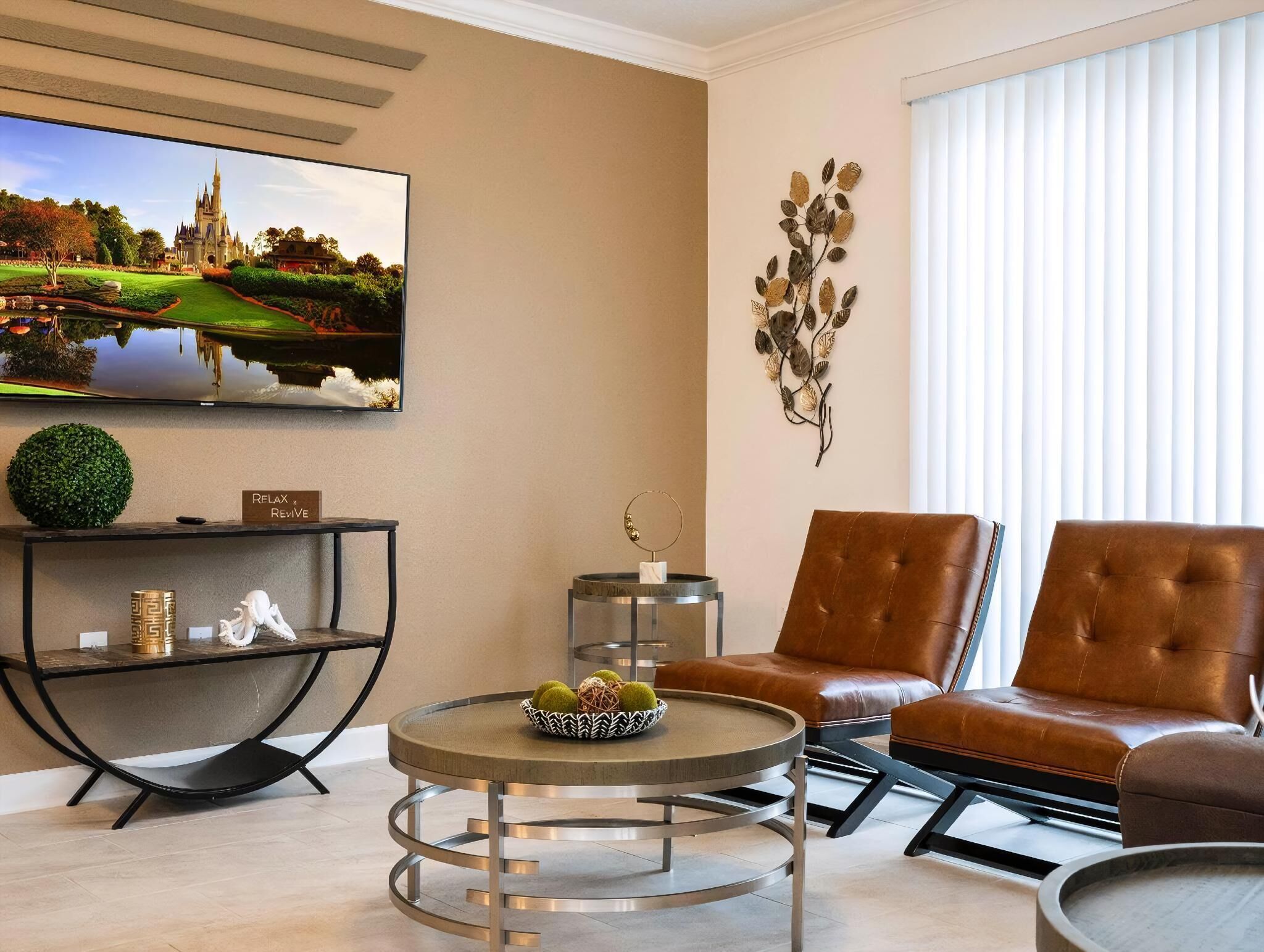 A modern living room with a round coffee table, two brown leather armchairs, a TV mounted on the wall, and a decorative wall piece.