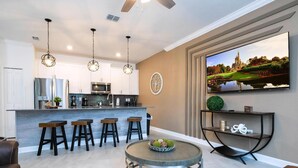 A modern kitchen with white cabinets, stainless steel appliances, and a bar area with stools. There's a round table with a decorative bowl in the living area, and a TV mounted on the wall displaying a scenic view. - Storey Lake Resort, Private Pool, Near Famous Attractions!! (Kissimmee)
