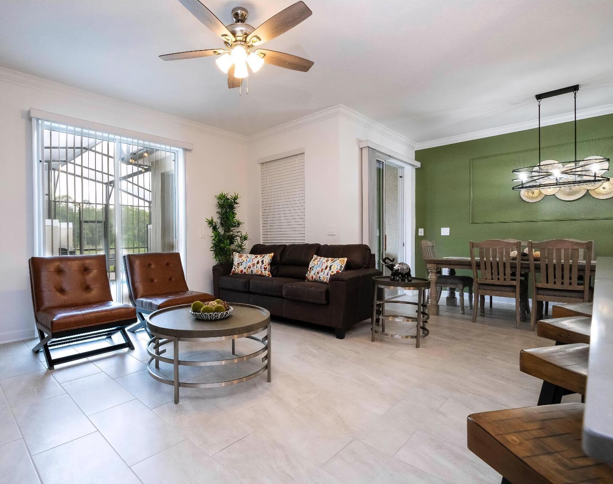 A spacious living room with a ceiling fan, a brown leather sofa, a round coffee table, and a dining area with wooden chairs and a green accent wall.