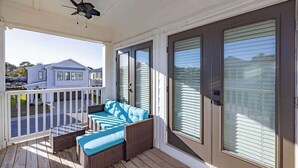 A covered porch with a ceiling fan, a wicker sofa with blue cushions, and a view of a blue house across the street. - Sleeps 16 - GAME ROOM, POOL, Perfect for Families (Orange Beach)