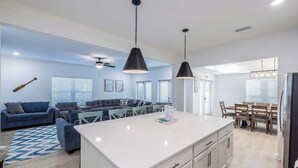 A modern kitchen with a central island, a dining area with wooden chairs, and a living room with a blue sofa and a baseball bat on the wall. - Sleeps 16 - GAME ROOM, POOL, Perfect for Families (Orange Beach)