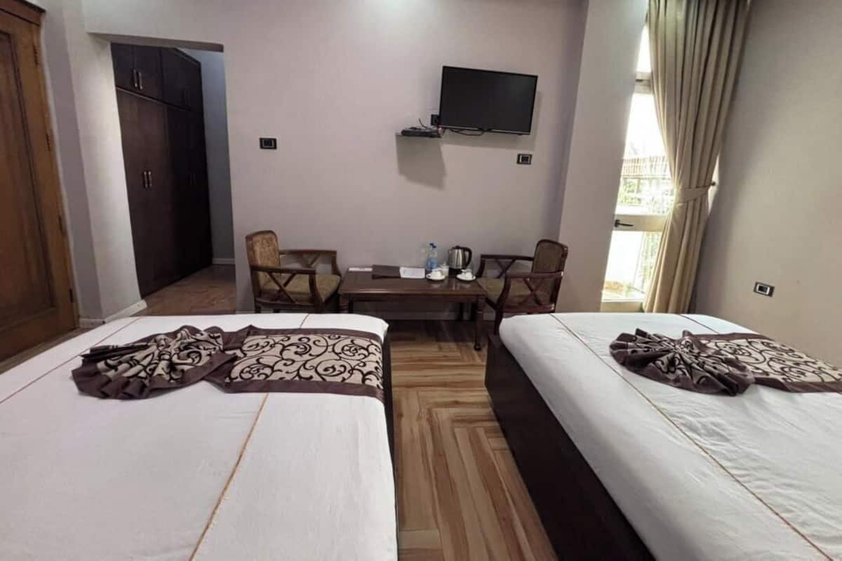 A hotel room with two beds, a TV mounted on the wall, a desk with chairs, and a window with curtains.