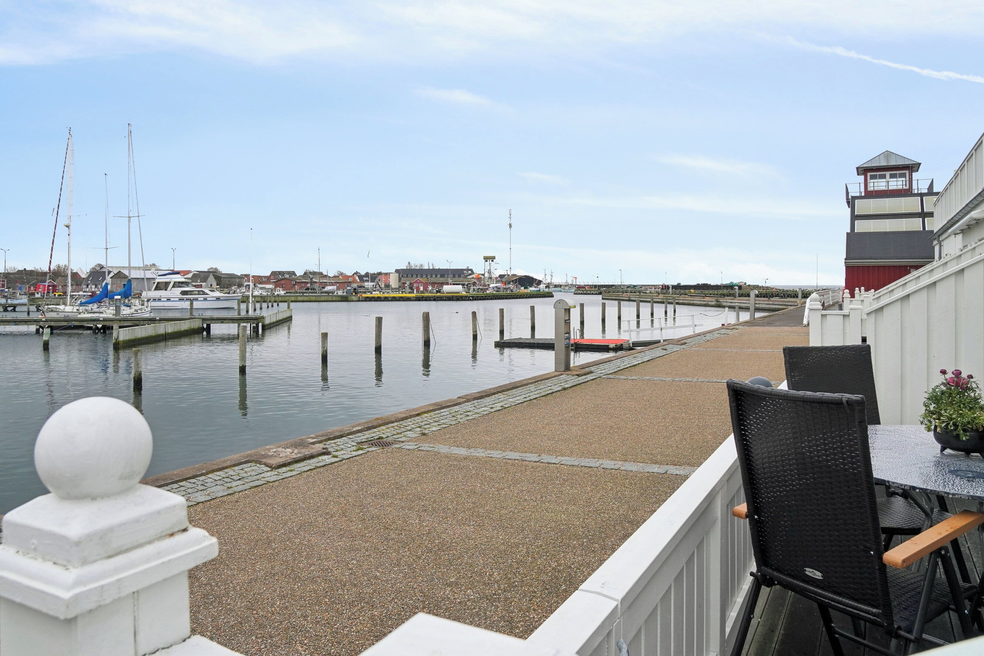 Um terraço à beira-mar com vista para o cais, um guarda-corpo branco e uma pequena mesa com cadeiras.