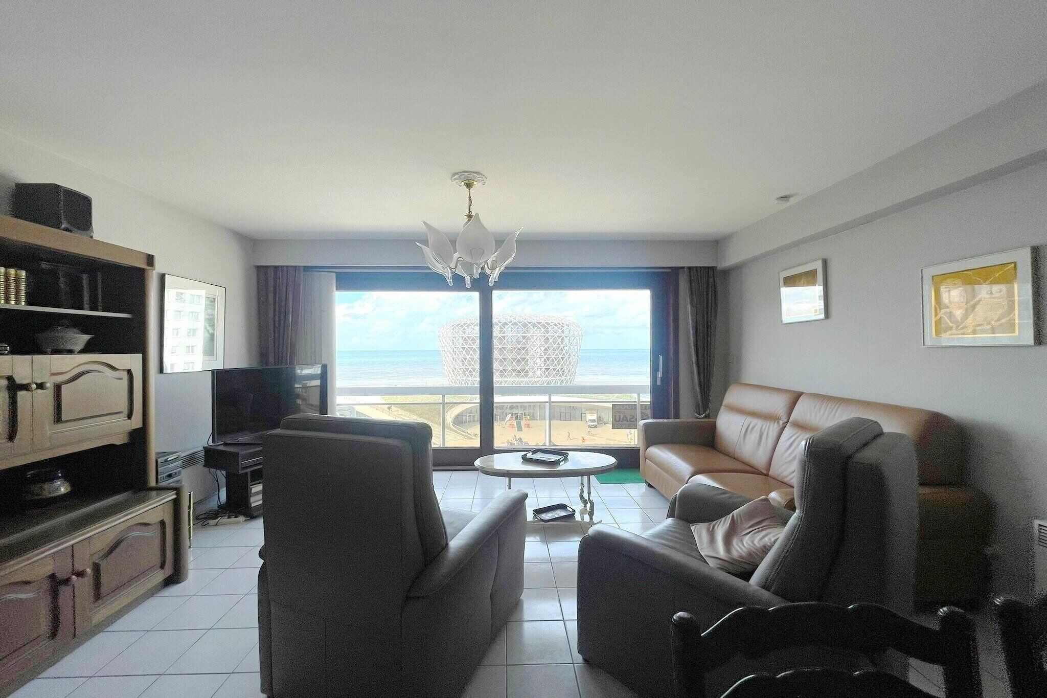 A modern living room with a sofa, armchairs, a coffee table, and a TV. A large window offers a view of the beach and a distinctive building.