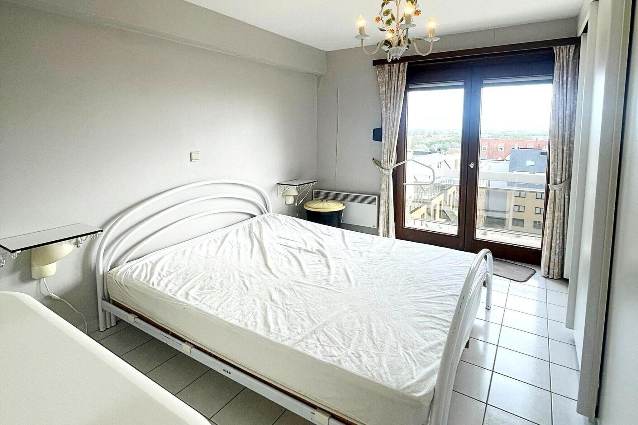 Une chambre à coucher comprenant un lit, une table de chevet, une lampe, une fenêtre avec des rideaux et une vue sur les bâtiments à l’extérieur.