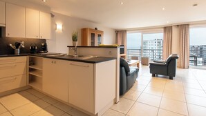 A modern kitchen with white cabinets, a black countertop, and a built-in island. There is a dining area with a black sofa and a chair, and a large window offering a city view. - Apartment in Middelkerke with Stunning Views (MIDDELKERKE)
