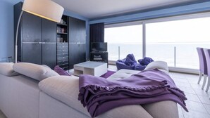 A modern living room with a sofa, a coffee table, a bookshelf, and a TV. There is a large window with a view of the sea. - Apartment Middelkerke with Sea View (MIDDELKERKE)
