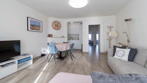 Una sala de estar moderna con un televisor de pantalla plana, un comedor con mantel rosa y un sofá gris con cojines decorativos.