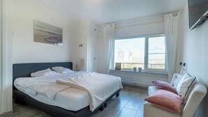 Un dormitorio con cama, una silla, un ventanal y un cuadro en la pared.