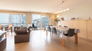 Een ruime woonkamer met een eettafel, banken en uitzicht op de stad door grote ramen.