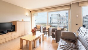 Een moderne woonkamer met een flatscreen televisie, een houten bijzettafeltje, een bank en een balkon met uitzicht op hoge gebouwen.
