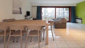 En moderne stue med et træspisebord, beige stole, en sofa og et stort vindue med udsigt til bygninger.