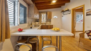 Een keuken met een centraal eiland, barstoelen, een eethoek met een rieten stoel en een houten plafond.