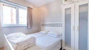 Un lit simple avec une literie blanche, une armoire blanche aux portes en verre et une fenêtre avec des rideaux.