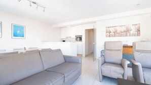 Un salon moderne avec un canapé et des fauteuils gris, une cuisine en arrière-plan et un coin repas avec une table et des chaises.