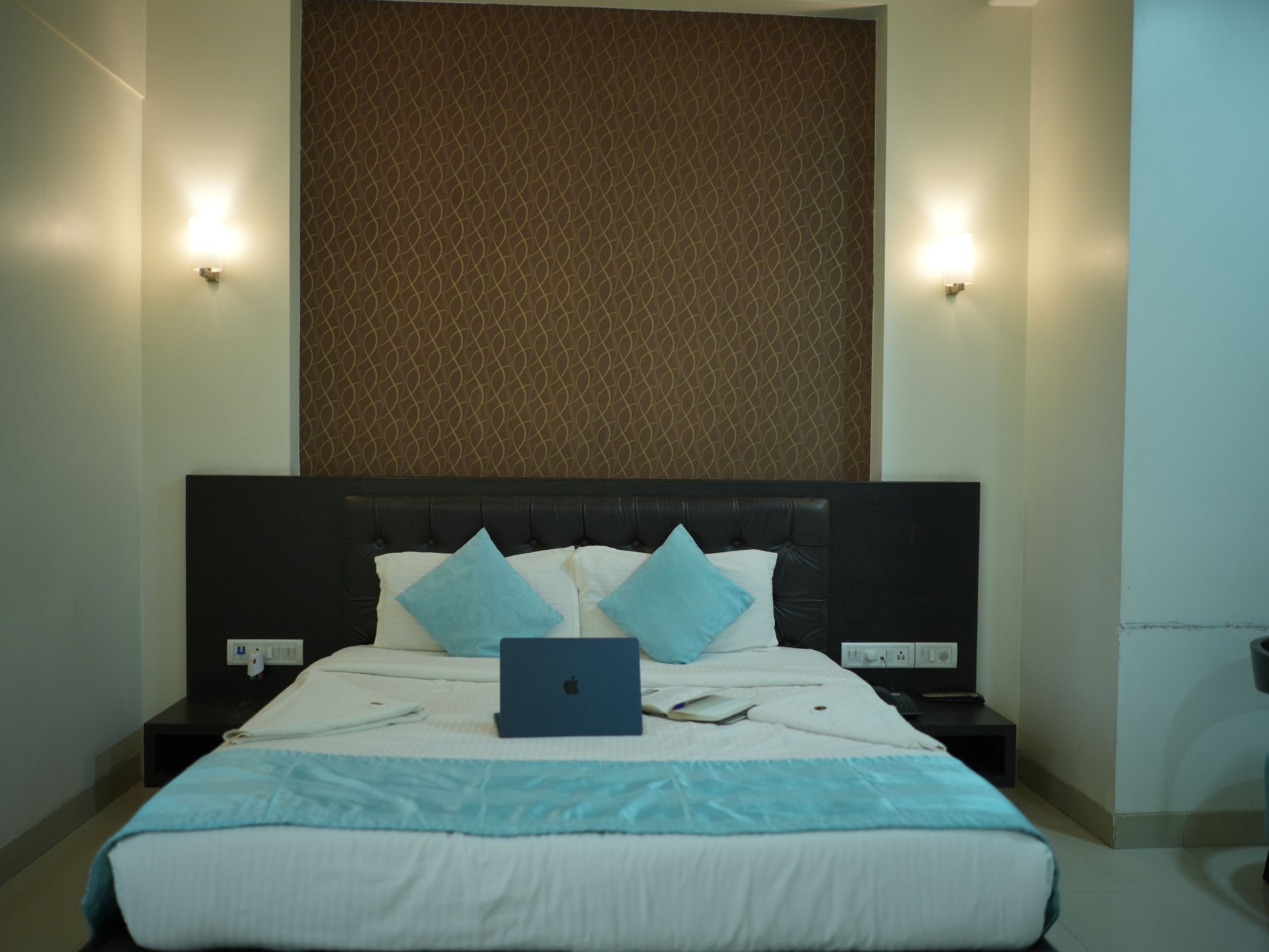 A neatly made bed with a laptop, pillows, and a blanket. There are two bedside tables with lamps and a wall-mounted light.