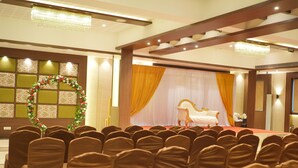 A banquet hall with rows of chairs covered in brown fabric, a stage with a white sofa, and a chandelier. - HOTEL WESTERN INN (Vasai)