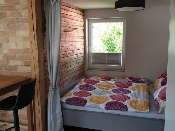 Una habitación acogedora con una cama, un ventanal con persianas, una pared de ladrillo y una mesa de madera.
