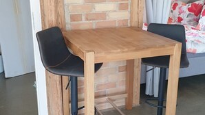 Una pequeña mesa de madera con dos sillas negras, un muro de ladrillo y una cama con ropa de cama floral al fondo.