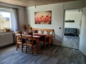 A dining area with a wooden table and chairs, a painting on the wall, and a sofa in the adjacent living room.