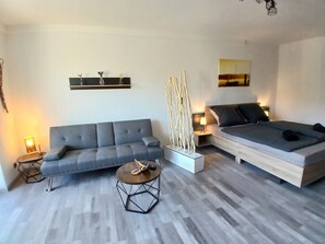 A modern living room with a grey sofa, a wooden coffee table, a decorative vase, and a bed with a grey mattress.