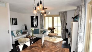 Ein modernes Wohnzimmer mit Esstisch aus Holz, weißem Sofa und dekorativer Beleuchtung.