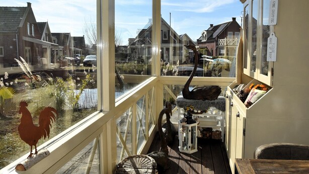Ein gemütliches Zimmer mit großem Fenster und Blick auf ein Dorf, einer Holzterrasse davor und rustikalem Interieur mit einer Laterne und Dekoration.
