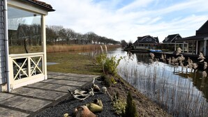 Ein Haus am Flussufer mit Wintergarten, einem Steinweg und einer Reihe von Häusern entlang des Wassers.