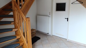 Una escalera de madera con peldaños negros, una puerta blanca con pomo negro, un radiador blanco y un felpudo negro en el suelo.