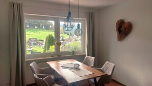 A dining area with a table, chairs, a hanging light fixture, and a view of a village through large windows.