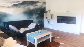 A living room with a black sofa, a wooden coffee table, a fireplace, and a wall mural of a forest. - Apartment 'Schöne Zeiten' with Mountain View, Private Terrace and Wi-Fi (Baiersbronn)