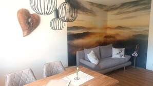 A dining area with a wooden table, grey upholstered sofa, and a wall mural depicting a sunset over a mountainous landscape.