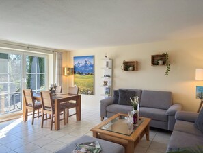 Un salon comprenant un canapé, une table basse avec un plateau en verre, une table à manger avec des chaises et une grande fenêtre.