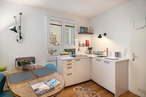 Une cuisine compacte avec des meubles blancs, une table à manger avec des chaises, une fenêtre avec des stores et une petite étagère avec des bocaux.