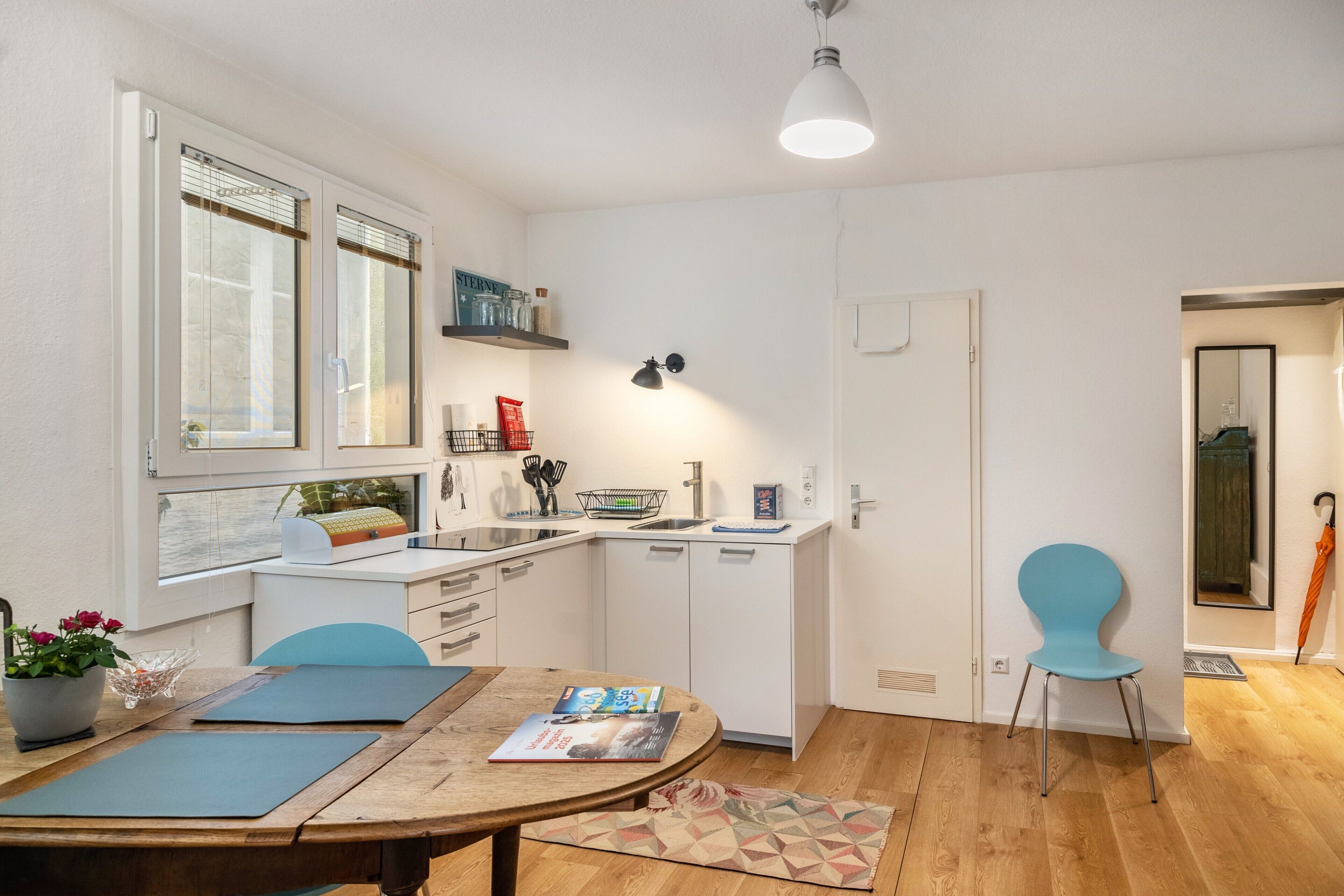 A compact kitchen with white cabinets, a dining table with chairs, and a small window with blinds.